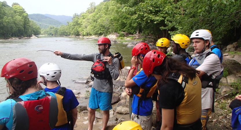 A group of students wearing safety gear watch as an instructor uses a stick to point out over a river.